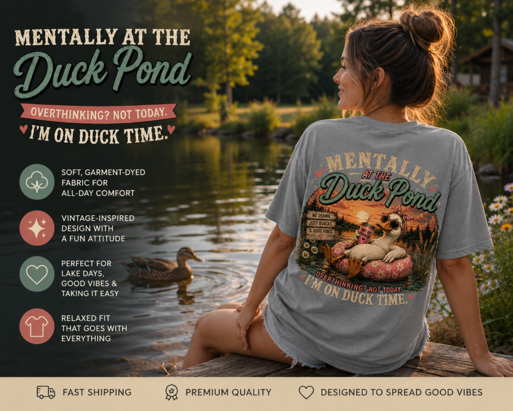 Woman wearing a gray “Mentally at the Duck Pond” graphic t-shirt sitting beside a peaceful pond at sunset, featuring a large vintage duck design on the back with relaxed lake-life vibes and rustic outdoor scenery.